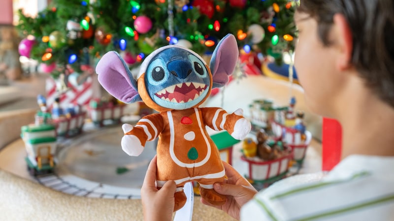 A boy holding a Stitch plush toy dressed in a gingerbread man costume near a Christmas tree