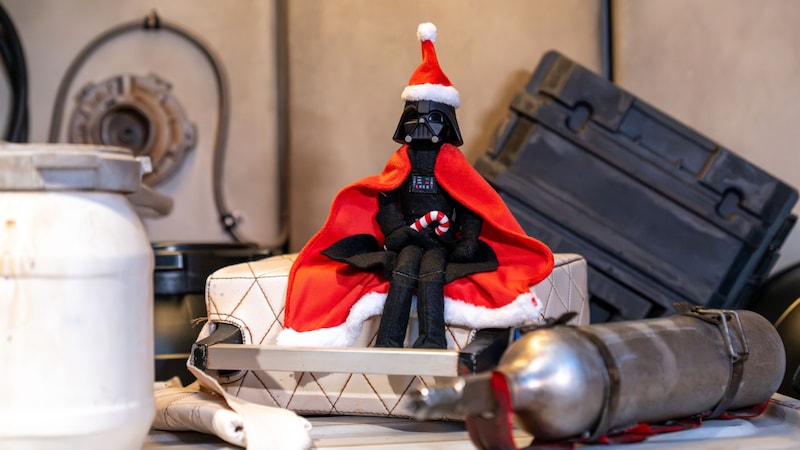 A Darth Vader plush wearing a Santa hat and holiday cape, holding a candy cane among weathered crates and mechanical props