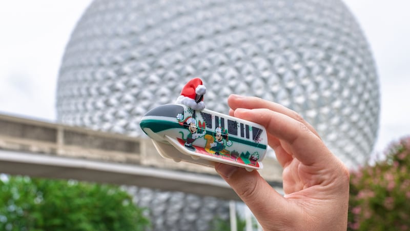 A hand holding up a small holiday themed Monorail toy decorated with a Santa hat near Spaceship Earth and the Monorail