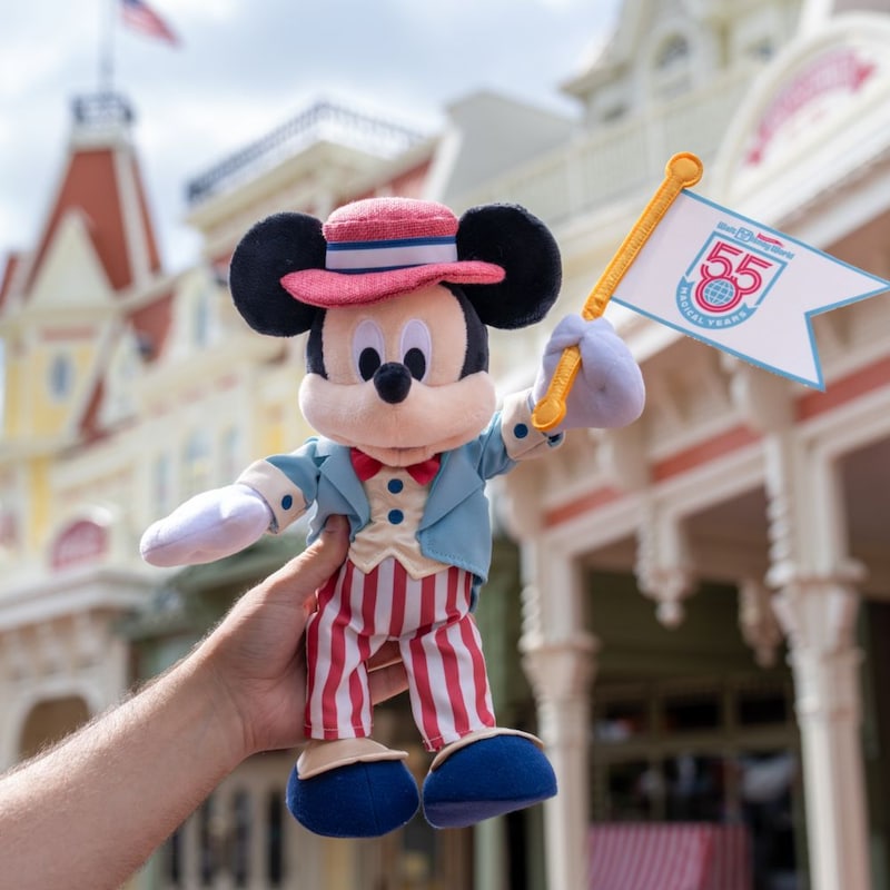 A Mickey Mouse plush dressed as a Main Street, USA performer holding a Walt Disney World 55th anniversary flag