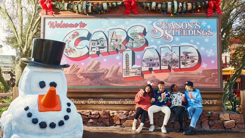 Mater made up as a snowman and young people enjoying snacks in front of a billboard reading ‘Welcome to Cars Land, Season’s Speedings’