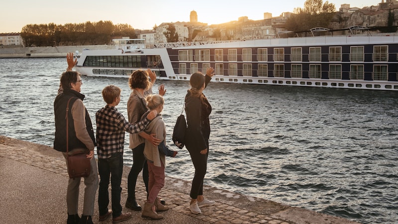 A family of five on land waves to a passing AmaWaterways ship