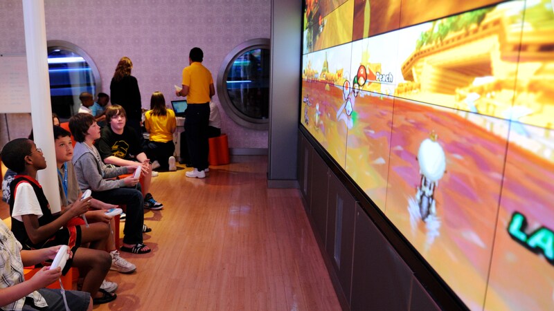 A row of kids sit, facing an enormous video screen with game controls in hand