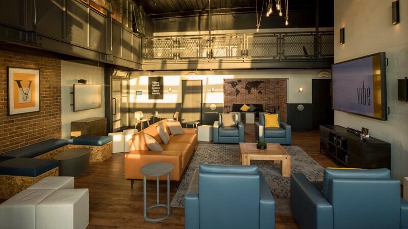 A loft style recreation room with a mix of modular seating, tables, wall art and 2 jumbo screen TVs