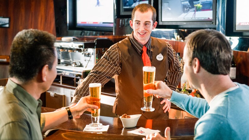A smiling Cast Member serves 2 tall glasses of beer to 2 men at a bar