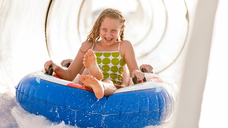 A joyful young girl holds on to the side handles of a raft as she waterslides through a translucent tube