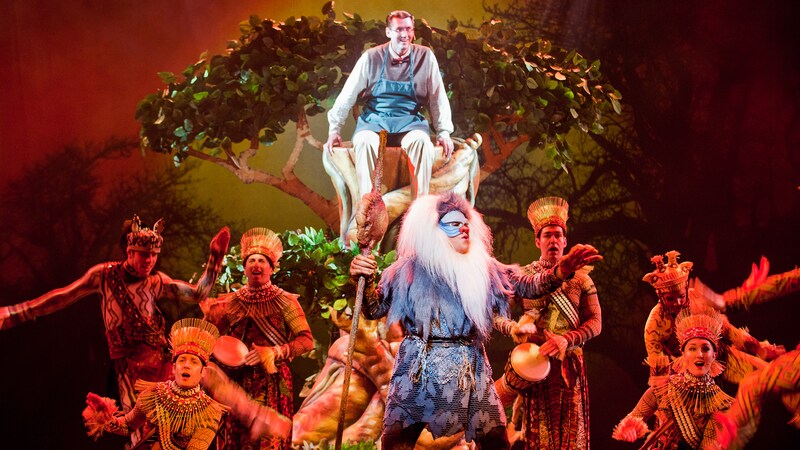 A father watches from a tree as Rafiki from The Lion King shares wisdom in a song accompanied by drums