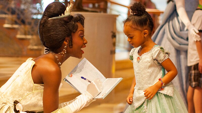 A small girl wearing a Tiana princess costume get an autograph from Princess Tiana