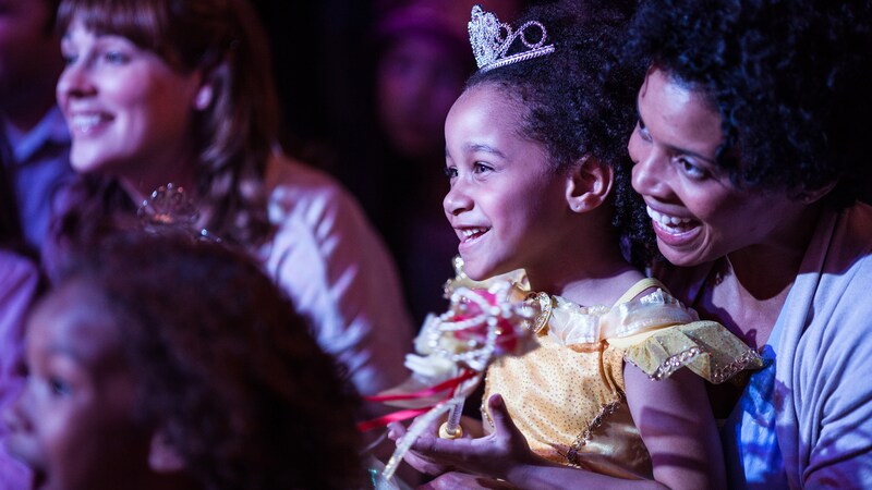 A smiling mother and her young daughter sit among other engaged audience members watching a live performance at the Walt Disney Theatre.
