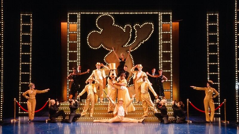Dancers in sparkling tuxedos and top hats pose on stage with Goofy at The Golden Mickeys