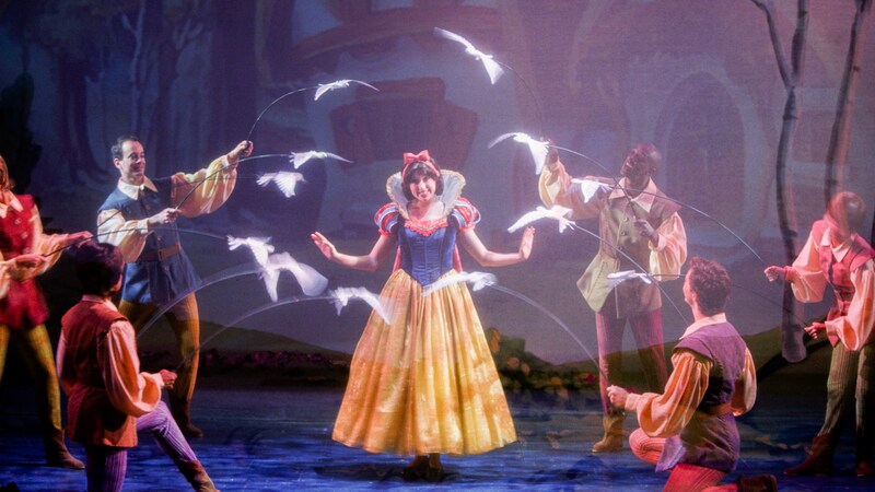 Birds dance in a circle around Snow White as she is met by villagers in a forest