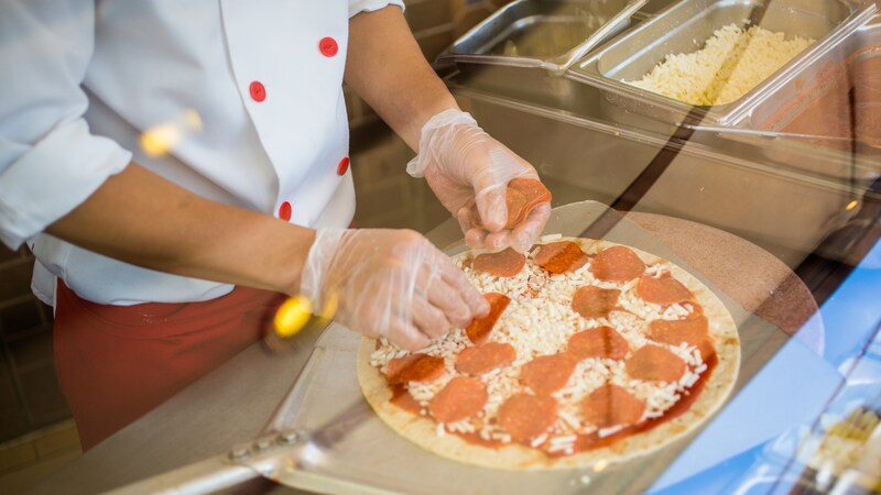 A pizzaria worker wearing plastic gloves prepares a pizza with pieces of pepperoni