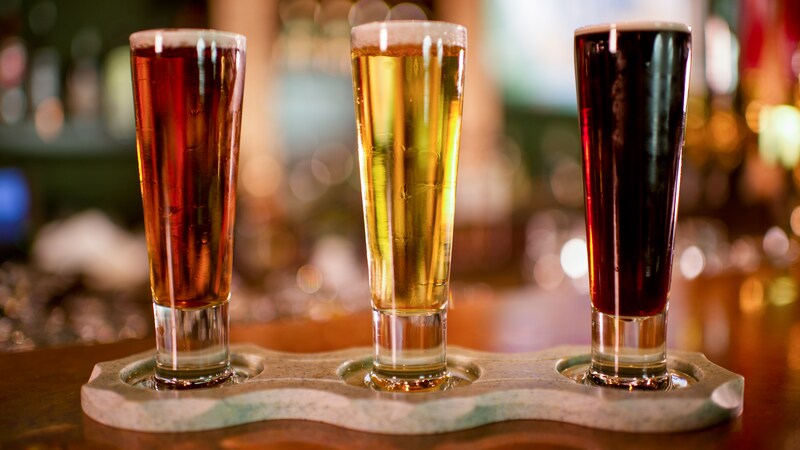 Three different types of beers in tall glasses are set on a 3 drink caddy on a bar