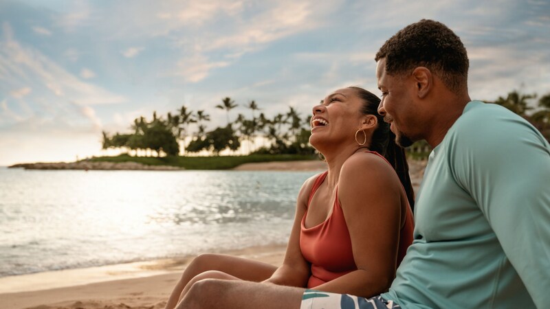 Two Guests sitting together on the beach while the sun sets over Aulani, A Disney Resort & Spa