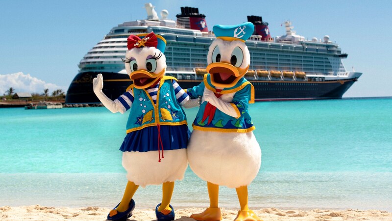 Daisy and Donald pose on a beach in their beachwear with a Disney Cruise Line ship in the background