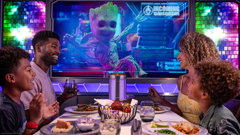 A young family of 4 enjoying a meal and an onscreen World of Marvel show featuring Baby Groot from Guardians of the Galaxy