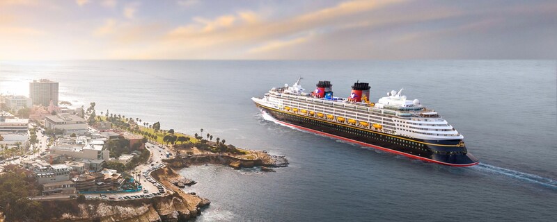 The cruise ship Disney Wonder sails near buildings along a rocky coast