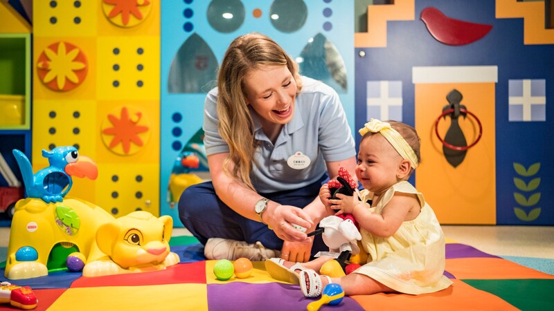 A female Crew Member sits on the floor with a laughing baby who is playing with toys