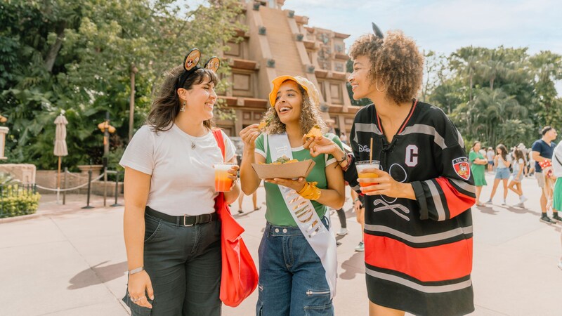 2 women with Minnie Mouse ear headbands enjoy cool, tropical drinks and share nachos with a friend