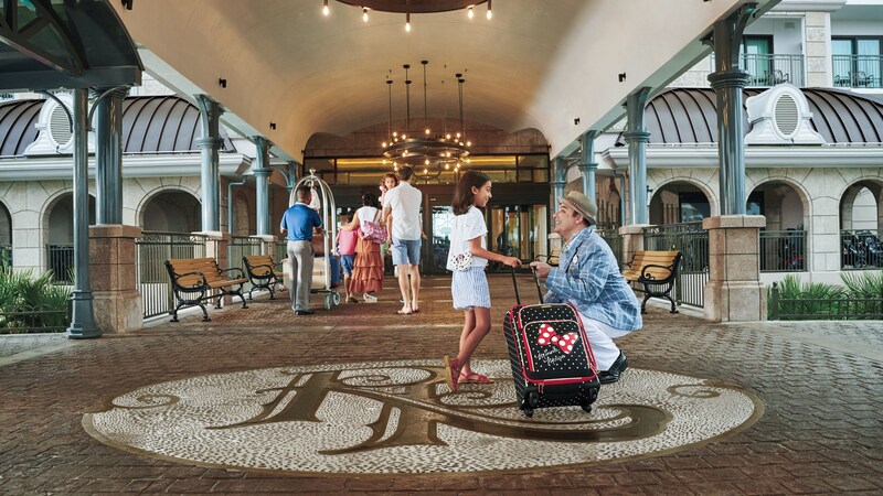 A Disney Cast Member greets a young girl who wheels her suitcase along the entry path in front of a Disney hotel