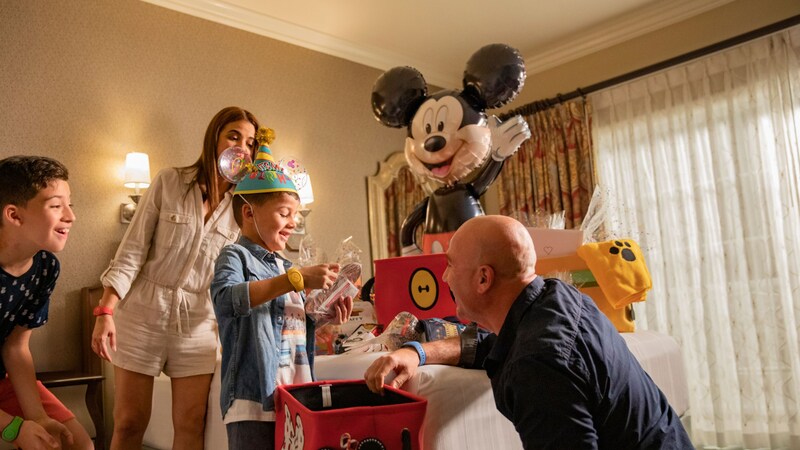 A family in a Disneyland Resort hotel room opening a Mickey Mouse birthday welcome basket featuring a Mickey Mouse ear hat and balloon 	