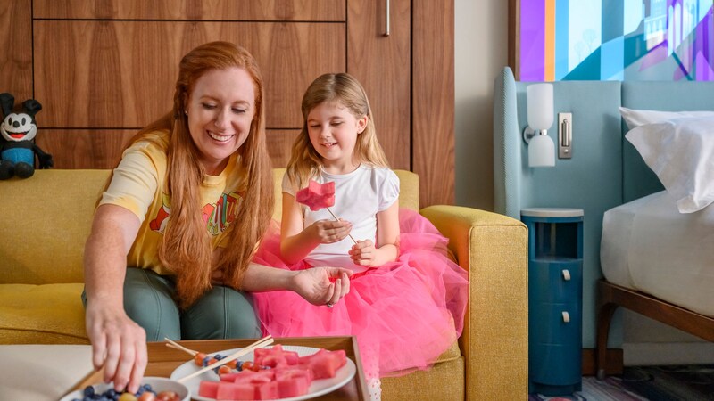 Mother and daughter enjoying a fruit snack together, sitting on a sofa at a Disneyland Resort hotel. 