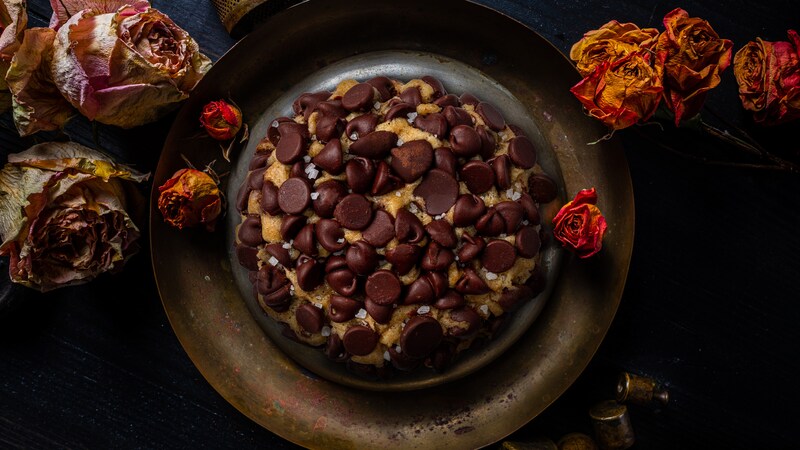 A chocolate chip cookie rests on a brass plate surrounded by dried roses