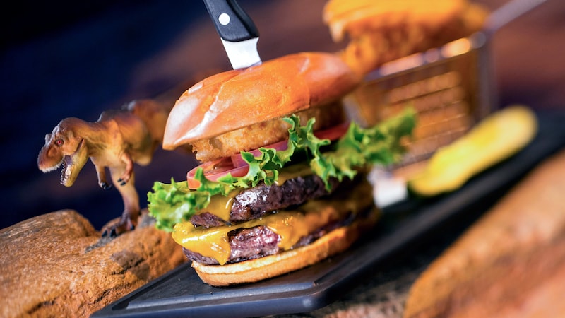 With a dinosaur figurine nearby, a double cheeseburger is topped with lettuce, tomato and an onion ring