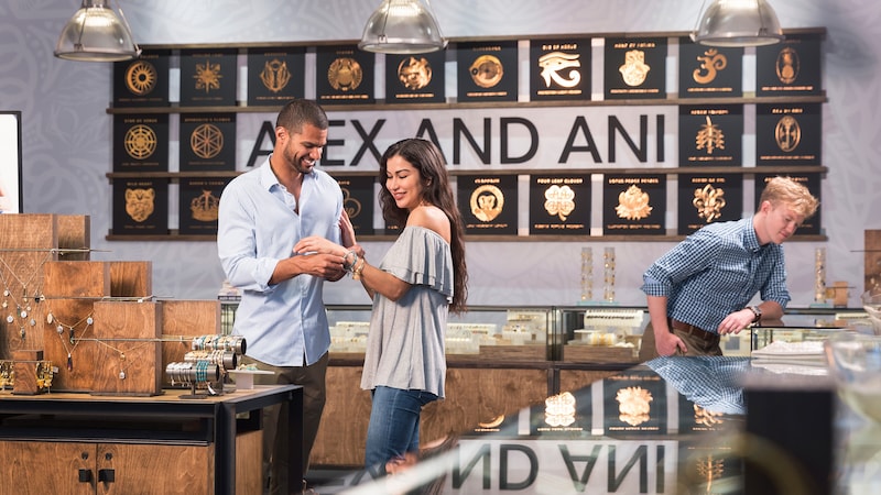 The ALEX and ANI store interior featues glass showcases holding jewelry as a man adjusts a bracelet on his girlfriends wrist 