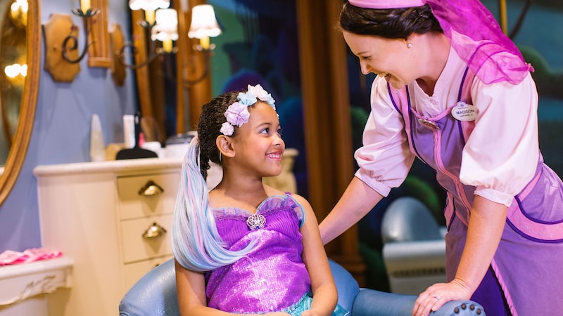 A Fairy Godmother talks to a seated little girl getting a princess makeover at Bibbidi Bobbidi Boutique