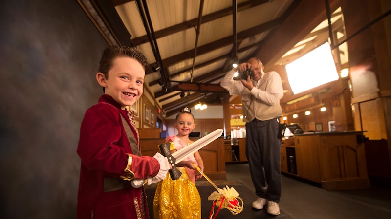 A Disney PhotoPass photographer holds a camera in studio with a boy dressed like a prince and girl dressed like Belle