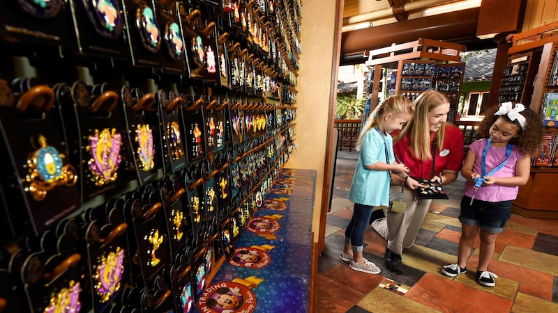 A Cast Member helps 2 little girls pick out pins next to an expansive wall display of pins