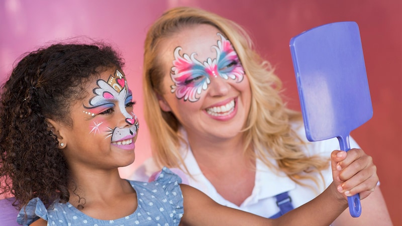 A young girl holds up a mirror to inspect her makeup at the Face Painting by Enjoy Your Face kiosk at Disney Springs as her face painting artist looks on