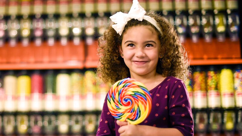 A smiling little girl holds a large lollipop inside Goofy’s Candy Company