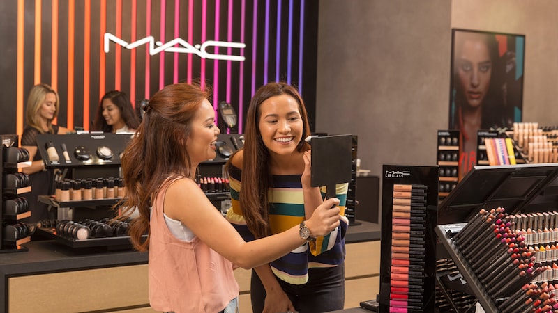 A young woman holds up a mirror for her female friend who is trying on makeup at MAC Cosmetics