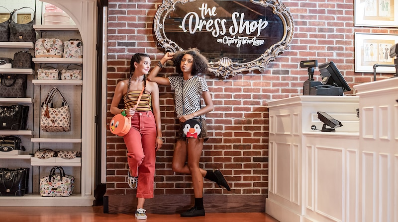 Two young women holding Disney themed bags stand against a brick wall near store displays and a checkout counter