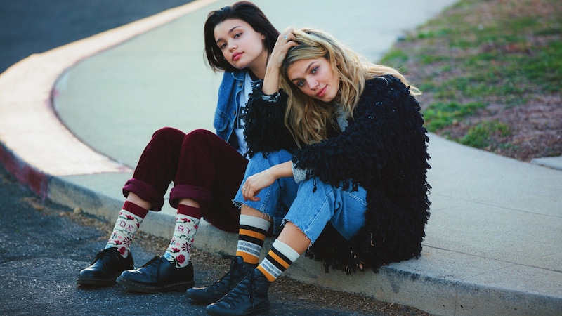 Two young females sit on a curb wearing stylish outfits with vibrant Stance socks