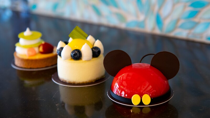 Three delightful circle shaped pastries, including one that looks like Mickey Ears, one cheesecake with whipped cream and fruit toppings, and one tart topped high with fruit and whipped cream