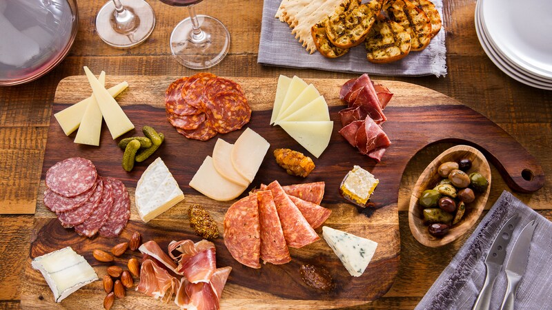A table with wine, plates, a bowl of olives, cheese knives and a charcuterie board of cheese and meats