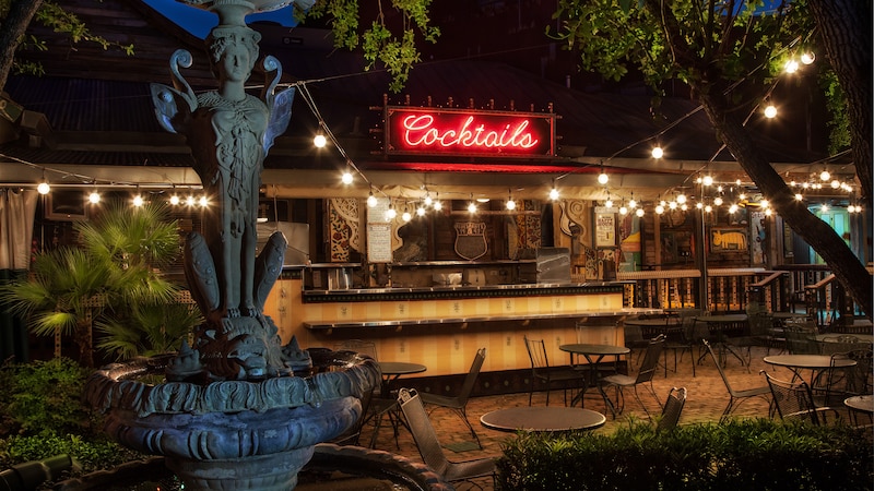 Sphinx-like fountain near patio tables and bar lit up with neon 'Cocktails' sign