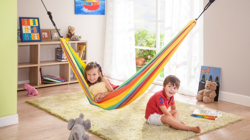 A girl sits in a hammock next to a boy in a sunny room