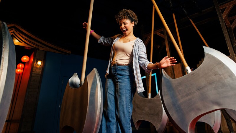 A young woman laughing while navigating between large swinging axe like pendulums in a darkened challenge room