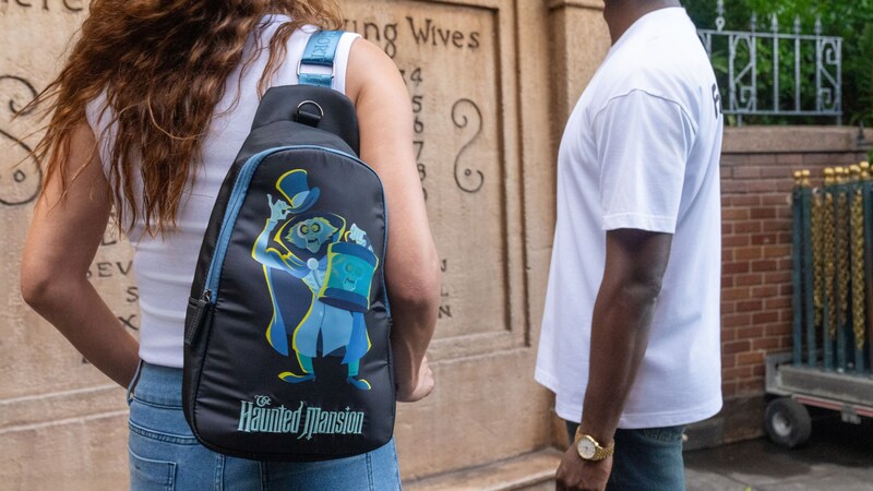 Dos visitantes de pie cerca de The Haunted Mansion, mientras uno de ellos lleva una mochila temática de Haunted Mansion.