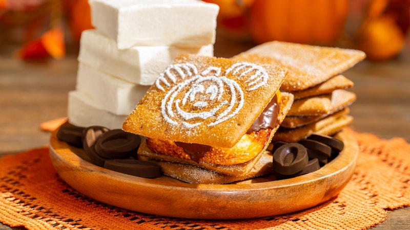 A pumpkin spice smores sandwich featuring Mickey Mouse plated next to marshmallows, chocolate and graham crackers