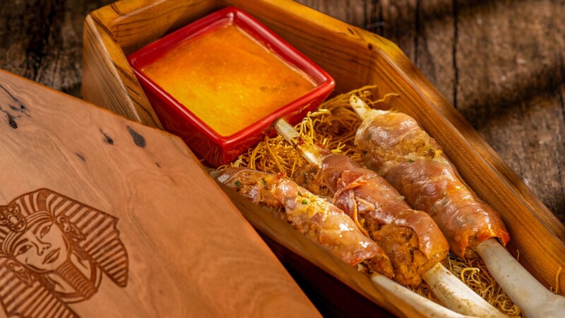 A wooden box resembling a coffin with pork and beef bone kabobs wrapped in prosciutto, served with tomato sauce