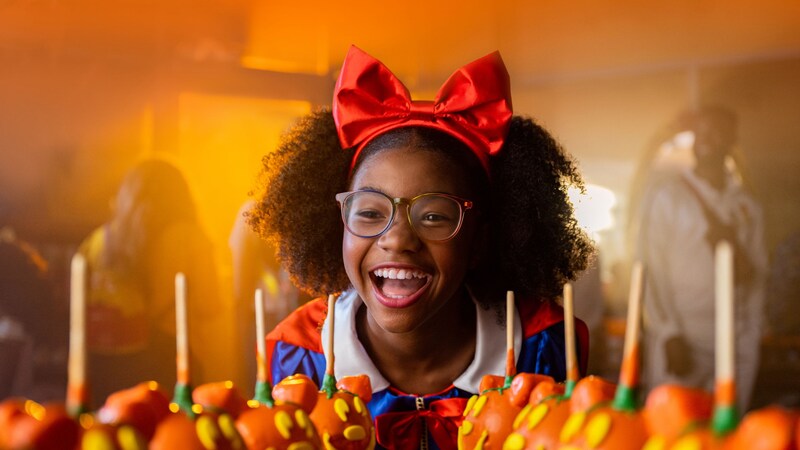 A young Guest smiles at 2 rows of caramel apples decorated like Mickey Mouse jack o lanterns