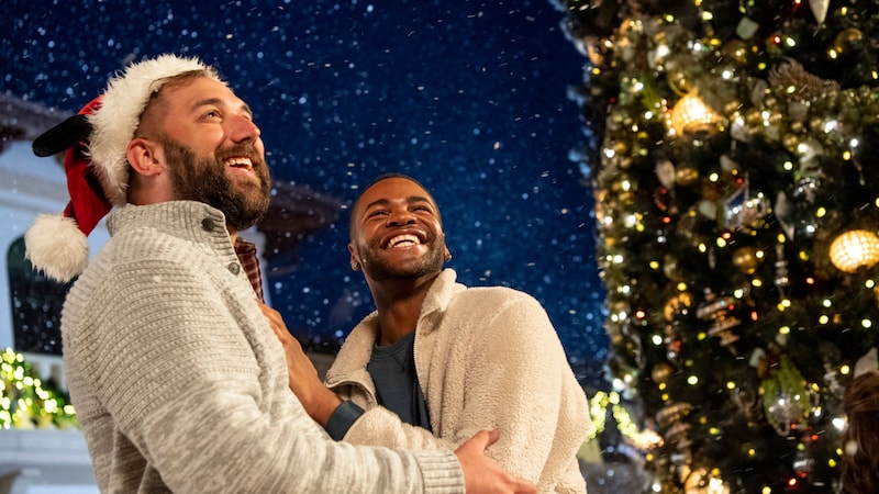 Two men hold each other and smile as snow falls near a Christmas tree