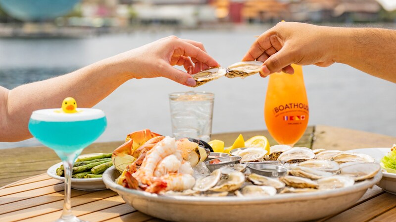 Two Guests toasting with oysters above a table filled with plates of oysters, prawns, asparagus and 2 cocktails at The Boathouse restaurant at Disney Springs
