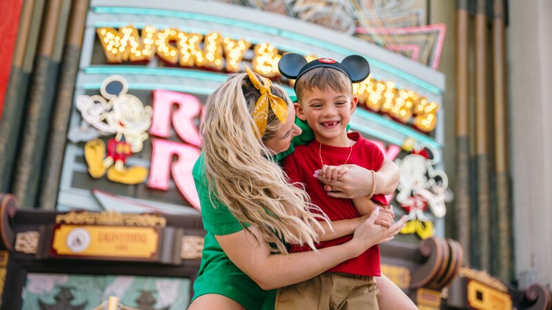 A boy in a Mickey Mouse ear hat gets a hug from his mom in front of Mickey and Minnie’s Runaway Railway