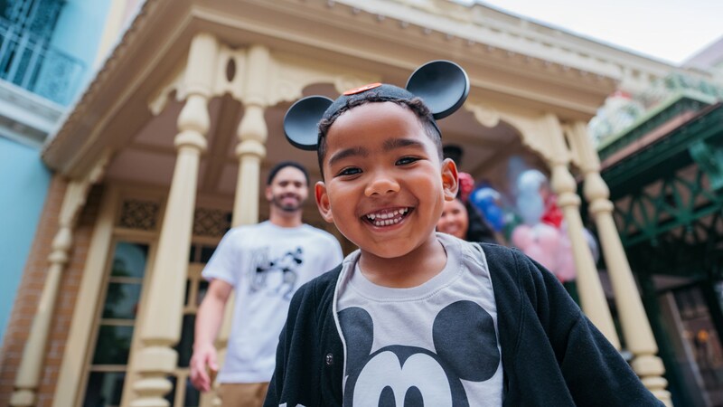 A young boy smiling and standing in front of his parents while wearing a Mickey Mouse ear hat and shirt
